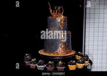 torta nera festiva a due piani con decorazioni in oro per il 30th° anniversario. molti cupcake al cioccolato e alla vaniglia con decorazioni di lusso. candybar per bi Foto Stock