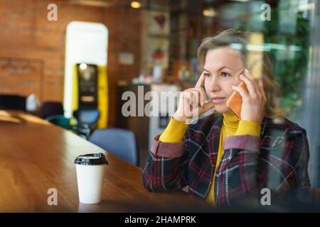 Foto di una splendida donna caucasica di mezza età seduta nel bar e parlare al telefono cellulare. Spazio per il testo. Messa a fuoco soft. Foto Stock