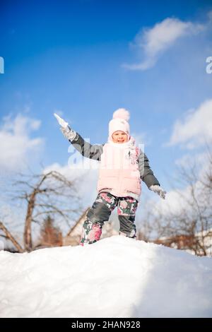 Il bambino si alza in cima alla nevicata. Bambina contro il cielo blu giocando palle di neve e saltando sullo scivolo di neve dopo una pesante tempesta di neve, paesaggio invernale Foto Stock