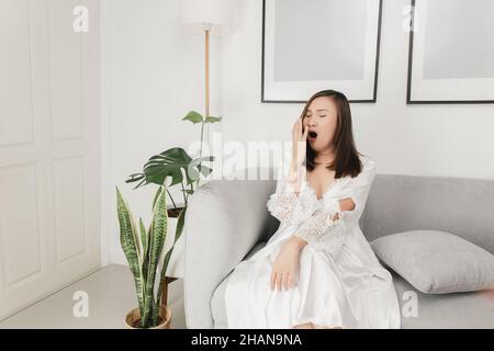 Donna thailandese in un nightgown satinato e vestito di abito bianco che si yawning sul divano nel soggiorno di notte Foto Stock