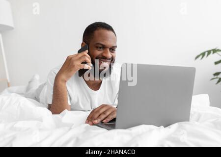 Sorridente giovane uomo nero con barba parla per telefono, guarda il computer portatile sul letto bianco nella camera da letto interna Foto Stock