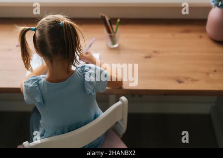 Vista posteriore ad angolo alto di poco non riconoscibile ragazza imparante scrittura facendo i compiti seduti al tavolo domestico dalla finestra. Foto Stock