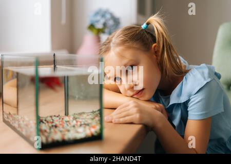 Graziosa ragazza caucasica che guarda il pesce d'oro in acquario, picchiettando sul vetro, attirando l'attenzione giacendo sul tavolo dalla finestra. Foto Stock