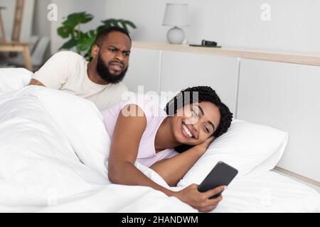 Triste giovane africano americano marito guarda il telefono della moglie sul letto bianco nella camera da letto interna Foto Stock