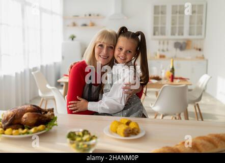 Ritratto di ragazza allegra e sua nonna in grembiuli che cucinano la cena di Natale, posando e abbracciando in cucina Foto Stock