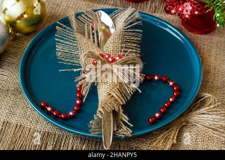 Elegante set di posate avvolto in tovagliolo di iuta su piastra di colore blu sulla superficie di tela con decorazioni natalizie Foto Stock