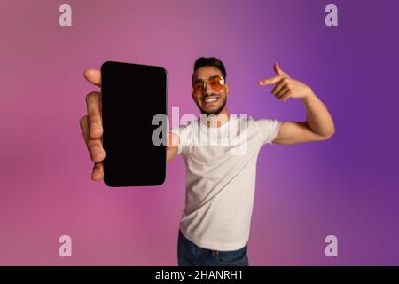 Giovane ragazzo arabo fresco che punta al cellulare con schermo nero vuoto, consigliando nuova app o sito web in neon luce, mockup Foto Stock