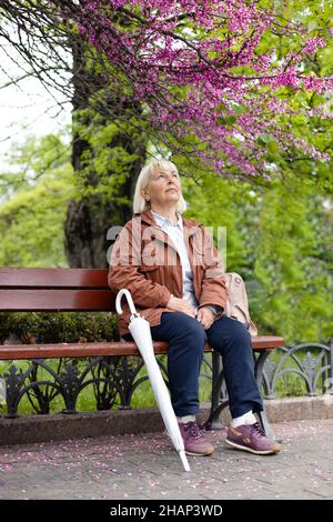 Felice caucasica 50s bionda donna con un ombrello trasparente si siede su una panca di legno in un parco estivo della città vicino ad un albero fiorente Foto Stock