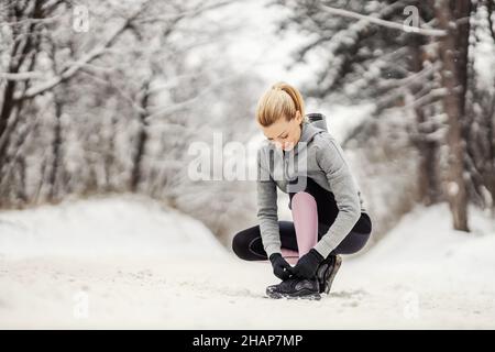 Sportswoman che si accovaccia su un sentiero innevato in natura e che lega le scarpe alla sua sneaker. Sportswear, vita sana, fitness invernale Foto Stock