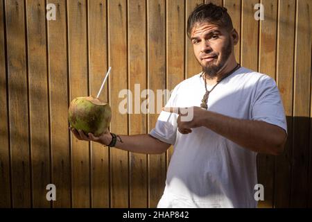 Uomo adulto con l'aspetto di un hippster o hippie e un cocco in mano. Atteggiamento comico che punta al frutto. Foto Stock