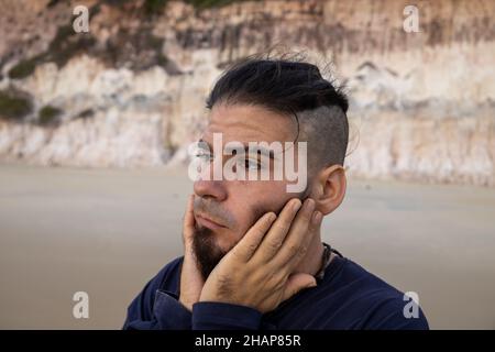 Giovane uomo di hippster o hippie aspetto con entrambe le mani tenendo il suo volto in preoccupazione sulla spiaggia Foto Stock