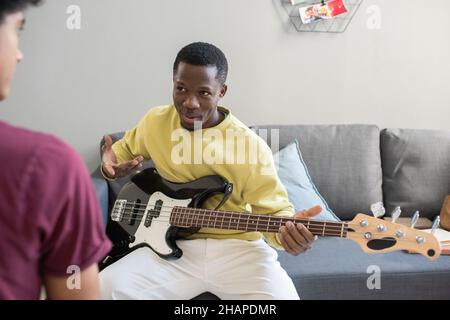 L'uomo africano in casualwear spiega agli studenti come suonare la chitarra mentre si siede sul divano a lezione di musica Foto Stock