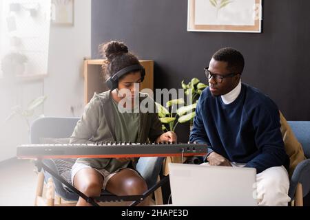 Carino diligente studentessa premendo i tasti della tastiera musicale mentre si siede accanto al suo insegnante in ambiente domestico Foto Stock