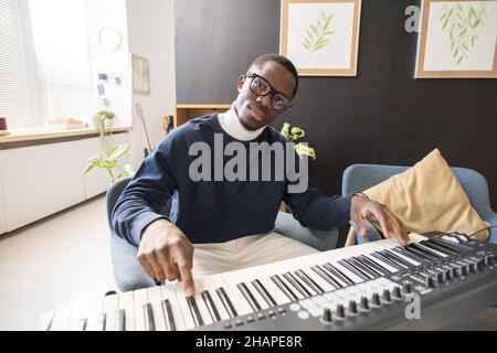 Giovane uomo africano serio che ti guarda mentre premi il tasto della tastiera musicale a lezione in ambiente domestico Foto Stock