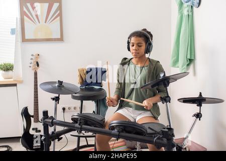 Ragazza giovane seria in cuffie seduto da drumkit di fronte alla fotocamera dello smartphone, mentre colpisce tamburi e cembali Foto Stock