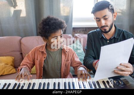 Carino ragazzino che guarda il foglio di musica tenuto dal suo insegnante che indica le note durante la lezione domestica di suonare il pianoforte Foto Stock