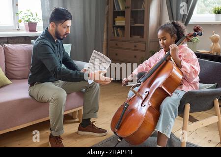 Carina studentessa che suona violoncello mentre la sua insegnante di musica si siede vicino e spiega come suonare lo strumento musicale Foto Stock