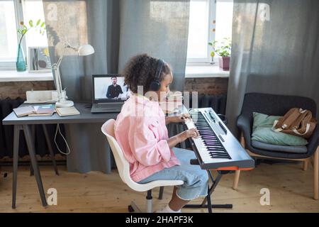 Vista laterale di una scolastica diligente che suona la tastiera del pianoforte mentre si siede contro la scrivania con il laptop durante la lezione online Foto Stock