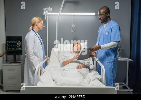 Donna anziana firma un accordo per l'operazione dando da parte dell'anestesista e medico in piedi vicino al letto in ospedale reparto Foto Stock