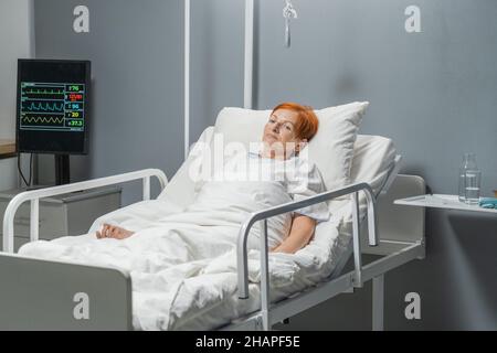 Donna anziana malata con espressione triste giacente nel letto d'ospedale dopo l'intervento Foto Stock
