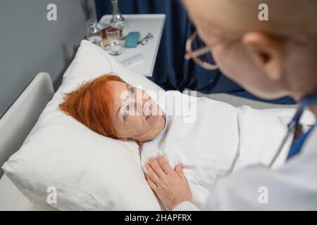 Donna anziana sdraiata a letto e guardando il medico che si prende cura di lei durante il trattamento in ospedale Foto Stock