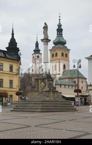 Colonna della peste nella piazza SNP a Banská Bystrica, Slovacchia. Il barbicano medievale con la torre dell'orologio è visto sullo sfondo a destra, mentre la Chiesa dell'Assunzione (Kostol Nanebovzatia Panny Márie) è vista a sinistra. Foto Stock