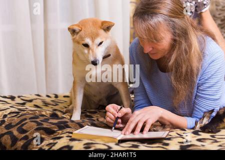 Una donna sdraiata a letto e scritta in un diario con il suo compagno cane shiba inu Foto Stock