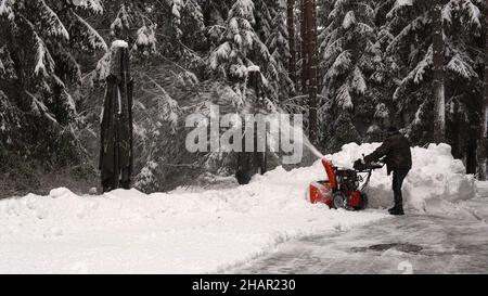 un uomo pulisce la neve con una spazzaneve sulla strada vicino a una foresta innevata invernale. Foto Stock