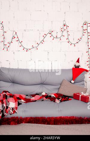 accogliente zona foto di natale. divano grigio con coperta a scacchi rossi. ghirlanda rossa su parete in mattoni bianchi. scatola regalo rossa e giocattolo babbo natale. Foto Stock