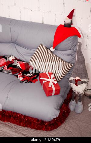 accogliente zona foto di natale. divano grigio con coperta a scacchi rossi. ghirlanda rossa su parete in mattoni bianchi. scatola regalo rossa e giocattolo babbo natale. Foto Stock