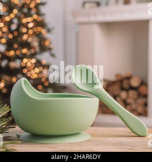 Set di capsule in silicone per bambini. Ciotola verde chiaro e cucchiaio sullo sfondo di albero di Natale e camino. Sfocatura creativa e grana. Inst. Telaio quadrato Foto Stock