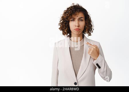 Donna d'affari sconvolta e poco sicura puntando il dito su se stessa e guardando insicuro, accigliando triste, in piedi su sfondo bianco Foto Stock