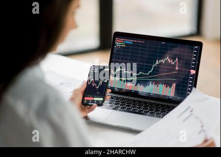 Un investitore e un operatore di crittografia di successo, utilizzando computer portatile e smartphone, analizza le carte di trading sul mercato azionario e lo scambio di criptovaluta digitale, conduce analisi, trading cripto monete Foto Stock