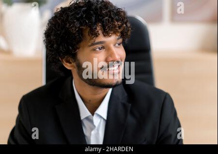 Headshot di attraente uomo d'affari positivo indiano o arabo, manager in abiti formali, si siede in ufficio, guardando positivamente al fianco, pensando alla startup o sognando una vacanza futura, sorridente Foto Stock