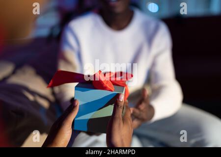 Le mani femminili africane che tengono la scatola del regalo avvolta, congratulandosi con il marito con anniversario in casa Foto Stock