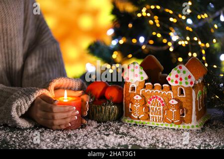 Bella casa di pan di zenzero e donna che tiene la candela a tavola contro sfondo sfocato Foto Stock