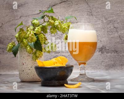 Bicchiere di birra con un rametto di luppolo e una ciotola con patatine su sfondo grigio. Foto Stock