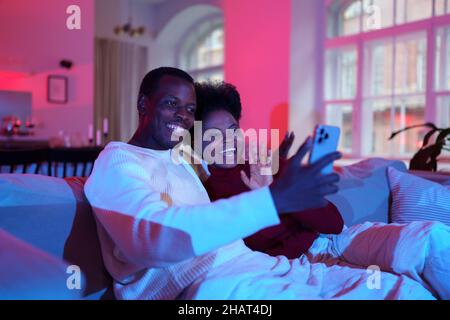 Allegra coppia africana parlare con gli amici tramite videochiamata su smartphone mentre si rilassa sul divano a casa Foto Stock