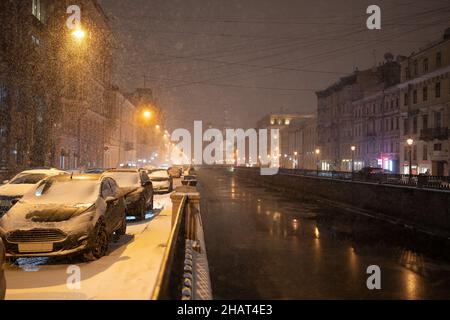 Strada invernale notturna vuota con auto parcheggiate lungo la strada durante le nevicate. Sera città in tempesta di neve Foto Stock