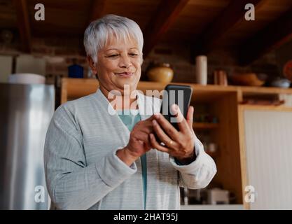 Contenuto multiculturale anziana femmina digitando un messaggio sullo smartphone mentre si trova in cucina moderna tenendo il caffè del mattino. Foto Stock