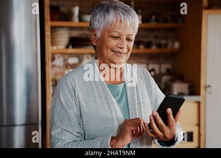 Sorridente anziana multiculturale che scrive un messaggio sullo smartphone mentre si trova in cucina moderna. Foto Stock