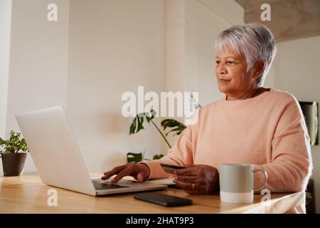 Anziana donna multietnica digitando nei dettagli bancari sul portatile, seduto al moderno banco cucina. Foto Stock