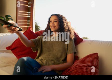 Multiculturale giovane adulta sorridente mentre sceglie cosa guardare in televisione. Appartamento moderno, comodamente seduto sul divano Foto Stock