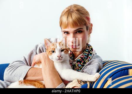 Soddisfare zenzero femmina seduta sul divano e abbracciando bianco e rosso gatto mentre guarda la macchina fotografica Foto Stock