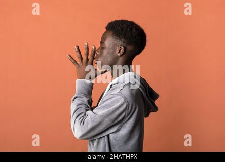 Vista laterale del giovane modello maschile africano americano con capelli ricci scuri in felpa con cappuccio grigio su sfondo arancione con occhi chiusi e. Foto Stock