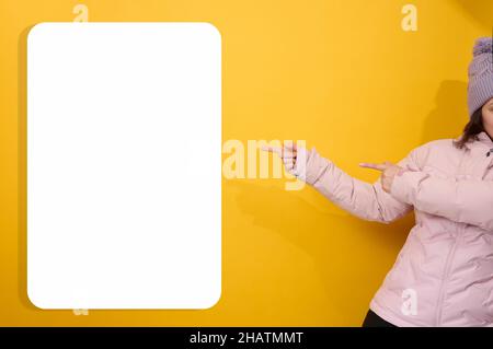 donna in una giacca rosa invernale e cappello mostra con le mani a lato su un cartellone bianco bianco bianco su sfondo giallo. Luogo per gli annunci, adv Foto Stock