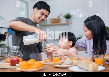 I genitori asiatici della famiglia thailandese si prendono cura del bambino giovane che mangia la colazione e beve il latte al mattino Foto Stock