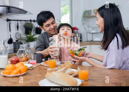 I genitori asiatici della famiglia thailandese si prendono cura del bambino giovane come mangiare la colazione di verdure e bere il latte al mattino Foto Stock