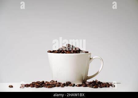 i chicchi di caffè fuoriusciti in tazza e sul tavolo su sfondo bianco. Caffè biologico in un mondo moderno minimalismo, dipendenza da caffeina. Vista frontale Foto Stock