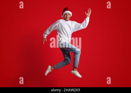 Divertimento per le vacanze. Giovane uomo allegro che indossa il cappello di Santa che salta su sfondo rosso Foto Stock